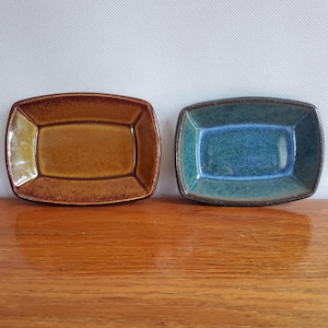 May include: Two rectangular ceramic dishes. One is a warm brown color, and the other is a deep teal. Both have rounded corners and a glossy finish. The dishes are displayed on a wooden surface.