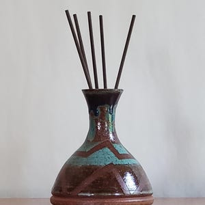 May include: A ceramic vase with a wide base and narrow neck, featuring a glazed finish in shades of brown and teal with a geometric zigzag pattern. Five thin, dark brown incense sticks are inserted into the opening of the vase.