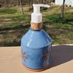 May include: A blue ceramic soap dispenser with a white pump and cork stopper. The dispenser has a speckled finish and is decorated with brown floral designs. The base is a lighter brown color.