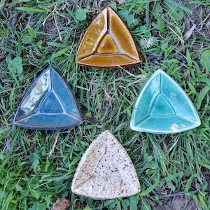 May include: Four triangular ceramic dishes arranged in a diamond pattern. One dish is glossy amber, one is deep blue with a speckled finish, one is a smooth light teal, and the fourth is a cream-colored ceramic with dark brown speckles throughout.