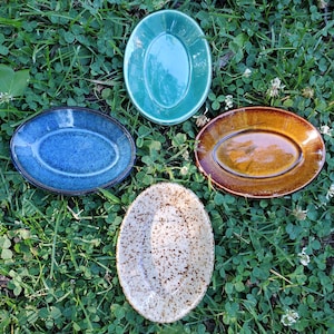 May include: Four oval-shaped ceramic dishes arranged in a cross pattern. The dishes feature distinct finishes: one glossy teal, one deep blue with a speckled texture, one warm amber brown with a smooth glaze, and one cream with dark brown speckles.