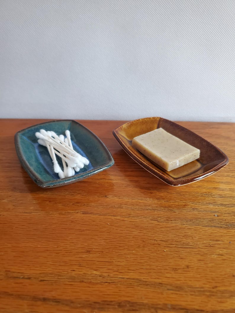 Może przedstawiać: Dwie prostokątne ceramiczne miseczki o zakrzywionych krawędziach. Jedna jest ciemnomorska i zawiera mały stosik białych patyczk&oacute;w higienicznych. Druga jest w ciepłym brązowym kolorze i zawiera prostokątną kostkę beżowego mydła z widocznymi drobinkami.