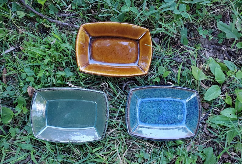 Może przedstawiać: Trzy prostokątne ceramiczne naczynia z podniesionymi krawędziami. Jedno naczynie jest błyszczące, bursztynowo-brązowe, drugie ma przygaszony szałwiowy odcień, a trzecie jest żywo niebieskie z nakrapianym wzorem. Każde ma gładkie, szkliwione wykończenie.