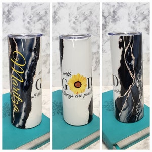 May include: A white tumbler with a black and white marble design and gold accents. The tumbler has the text "with God all things are possible" and a yellow sunflower graphic. The tumbler is sitting on a blue book.