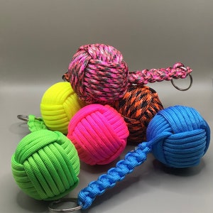 May include: A set of six colorful knotted rope balls with keychains. The balls are in various colors, including pink, green, blue, yellow, and black. The balls are made of braided rope and have a knot at the top. The keychains are attached to the balls with a metal ring.