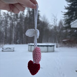 May include: A handmade crochet ornament featuring a red heart, a pink and white oval shape, and a white cloud-like shape, all connected by a white string. The ornament is held up against a snowy outdoor backdrop.