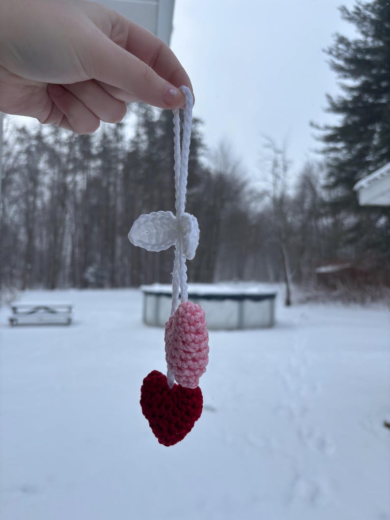 May include: A handmade crochet ornament featuring a red heart, a pink and white oval shape, and a white cloud-like shape, all connected by a white string. The ornament is held up against a snowy outdoor backdrop.