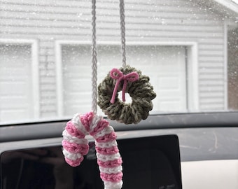 May include: Crocheted car charm with a candy cane and a wreath. The candy cane is white and pink, the wreath is green with a pink bow. Both are suspended by white cords, adding a festive touch to the car's interior.