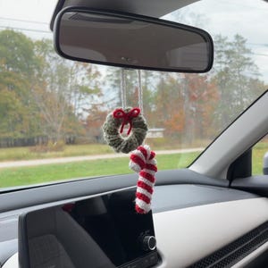 May include: A festive car charm, comprising a small, crocheted green wreath with a red bow and a red and white striped candy cane. The decorations are suspended from a car's rearview mirror.