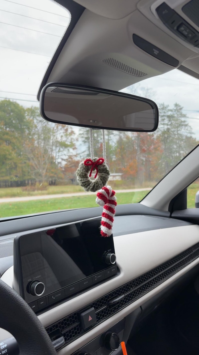 May include: A festive car charm, comprising a small, crocheted green wreath with a red bow and a red and white striped candy cane. The decorations are suspended from a car's rearview mirror.