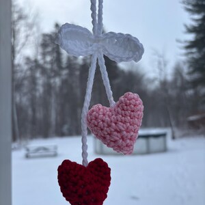 May include: A handmade crochet heart ornament. The ornament features a white bow and string, with a pink and a red heart hanging below. The hearts are made of yarn and have a textured appearance. The background is a snowy outdoor scene.