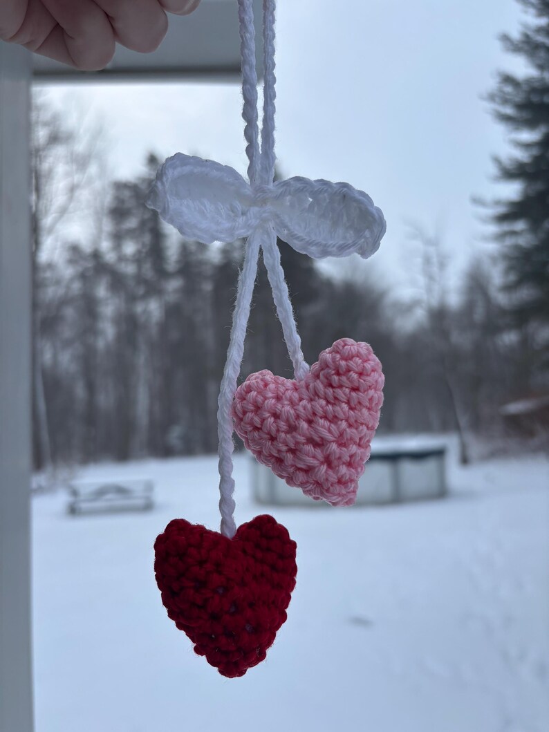 May include: A handmade crochet heart ornament. The ornament features a white bow and string, with a pink and a red heart hanging below. The hearts are made of yarn and have a textured appearance. The background is a snowy outdoor scene.