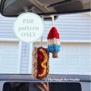 May include: Crocheted car charms in the shape of a hot dog and a popsicle, hanging from a car's rearview mirror. The hot dog has brown bread, red ketchup, and yellow mustard. The popsicle is red, white, and blue. Text on the image reads "PDF pattern ONLY".