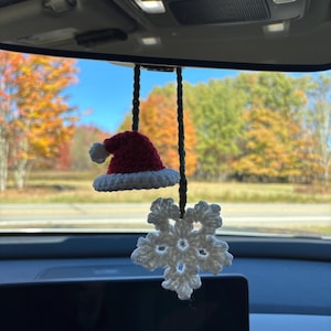 May include: A crocheted Christmas car charm featuring a red Santa hat with a white brim and a white snowflake. The charm hangs from a car's rearview mirror, with a scenic autumn background visible through the windshield.