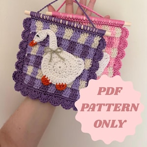 May include: Crocheted wall hanging featuring a white goose with orange feet and beak, a gray bow, and a purple and cream checkered background. The banner has a scalloped edge and is suspended by a wooden dowel and pink and purple yarn. The text "PDF PATTERN ONLY" is visible.
