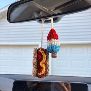 May include: Crocheted car charm featuring a hot dog with yellow mustard and a red, white, and blue popsicle. The charms are suspended from a rearview mirror. The hot dog is brown and the popsicle is red, white, and blue.