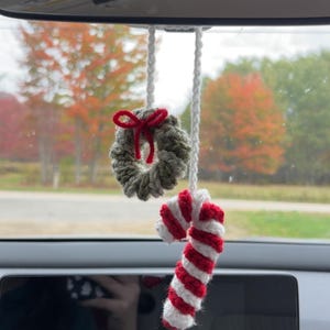 May include: Crocheted Christmas car charm featuring a small wreath with a red bow and a candy cane with red and white stripes. The ornaments hang from a white string, perfect for holiday decor.