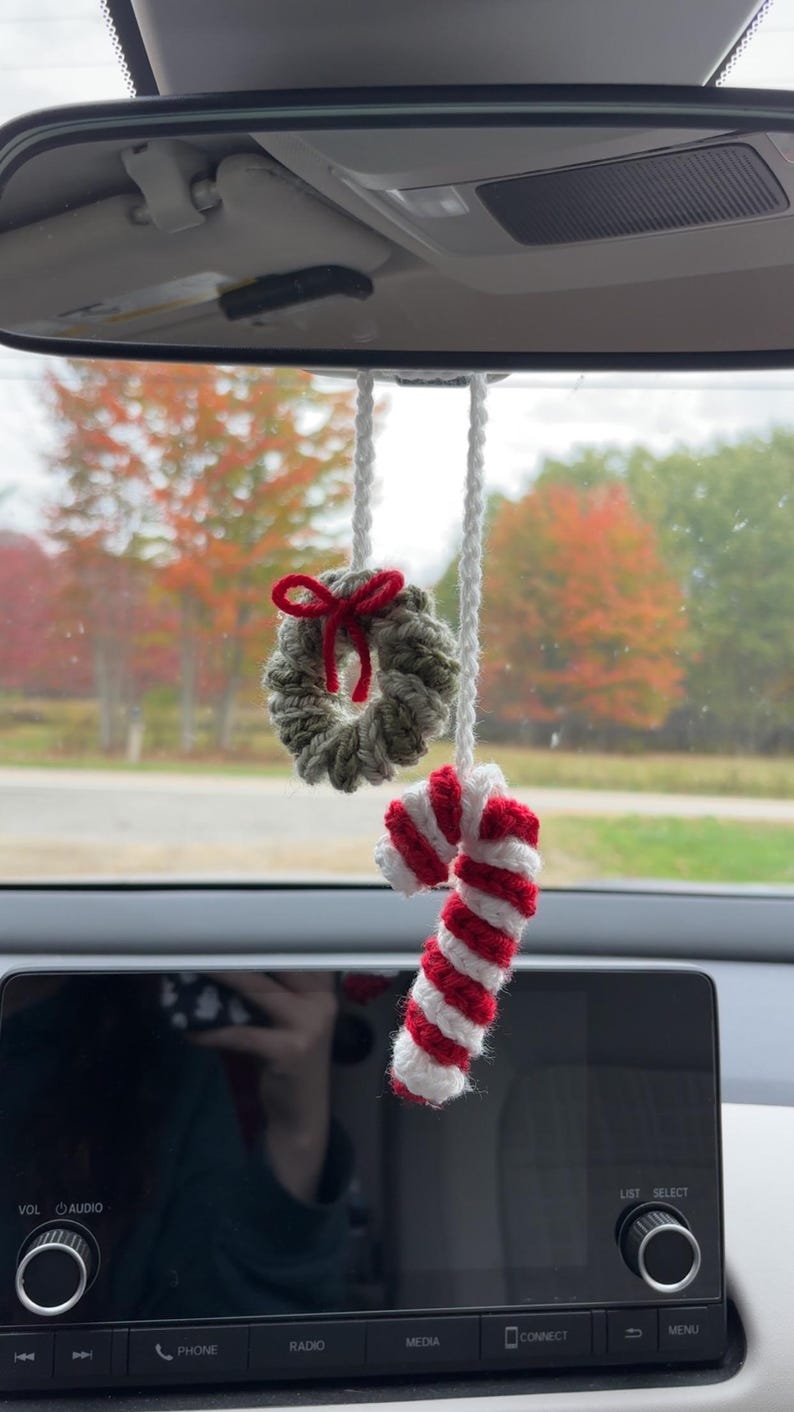 May include: Crocheted Christmas car charm featuring a small wreath with a red bow and a candy cane with red and white stripes. The ornaments hang from a white string, perfect for holiday decor.