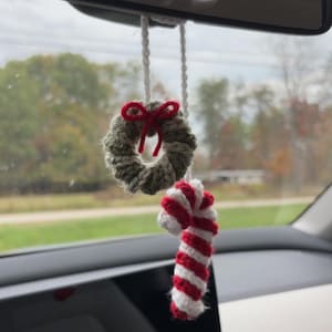 May include: A festive crocheted car charm. It features a small green wreath with a red bow and a red and white striped candy cane. The charm hangs from a white string, perfect for adding a touch of Christmas to a car.