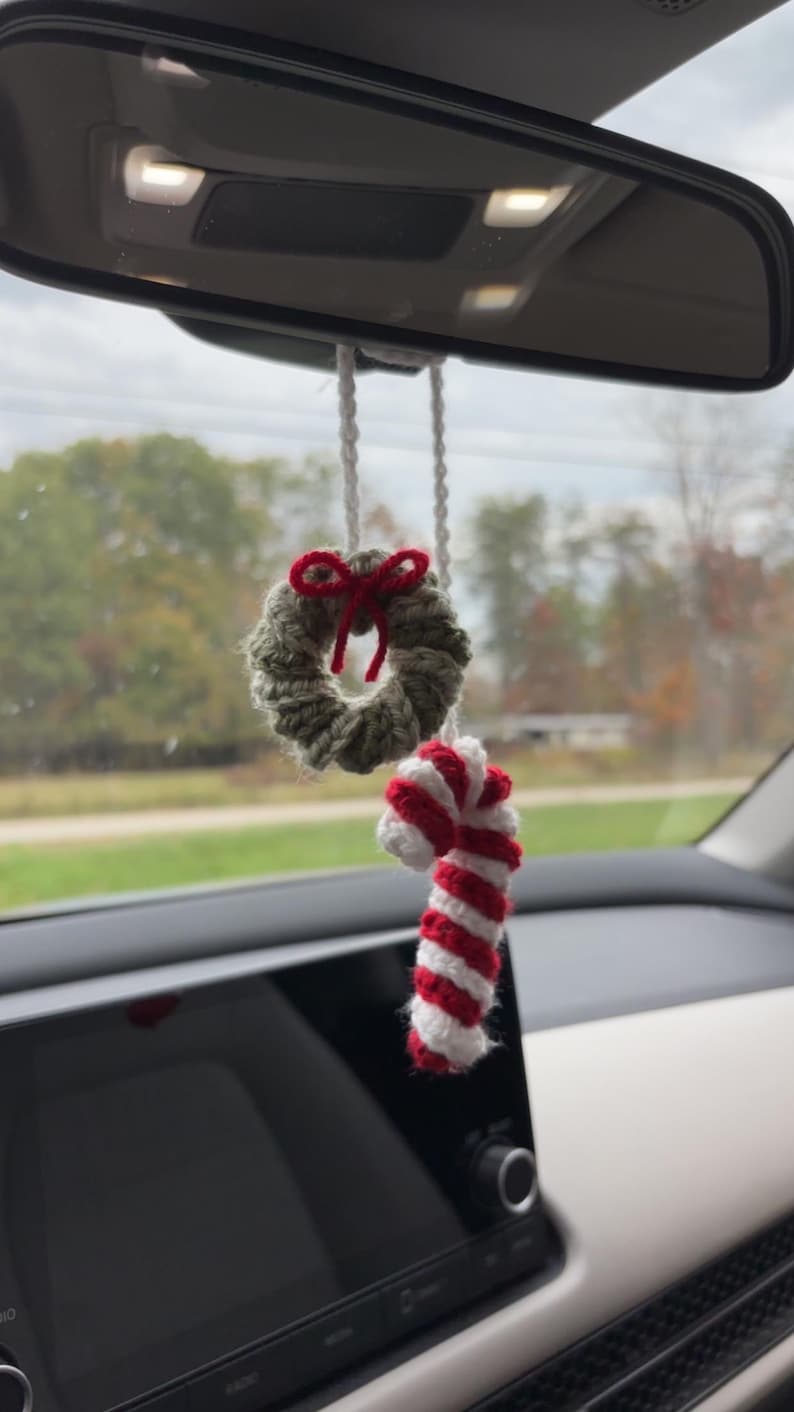 May include: A festive crocheted car charm. It features a small green wreath with a red bow and a red and white striped candy cane. The charm hangs from a white string, perfect for adding a touch of Christmas to a car.