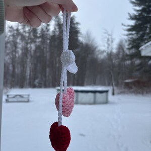 May include: A handmade crochet ornament featuring a white top element, a light pink heart, and a deep red heart. The ornament is suspended by a white cord. The background is a snowy outdoor scene.