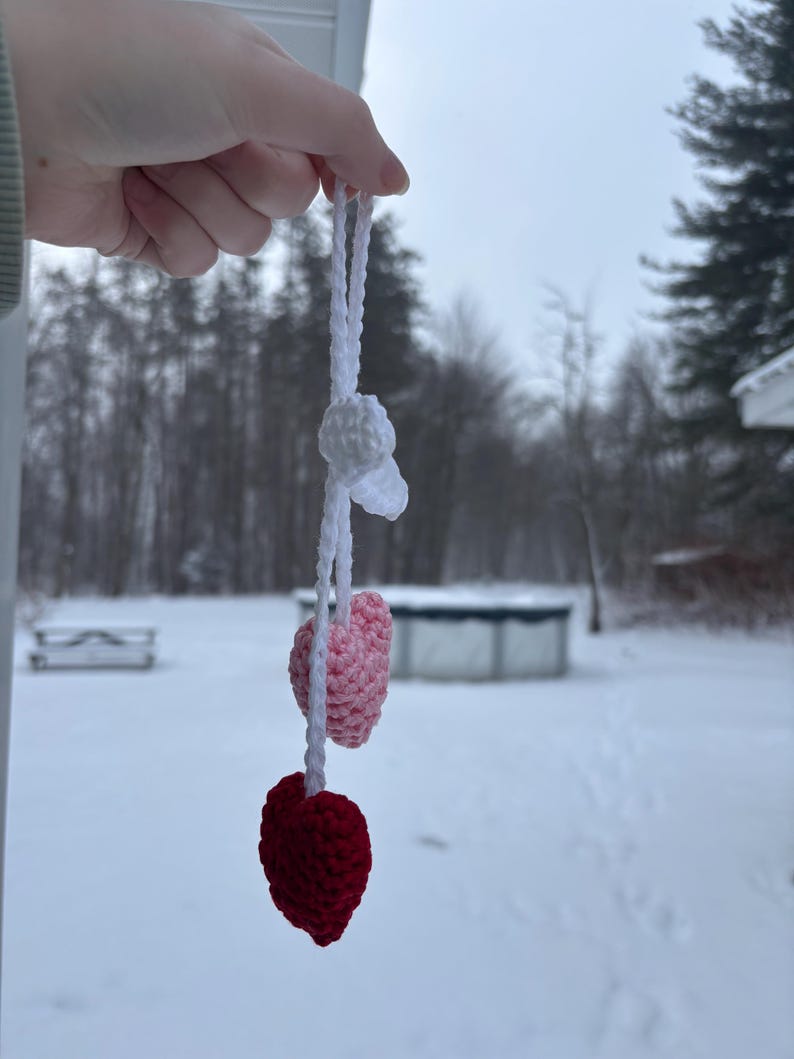 May include: A handmade crochet ornament featuring a white top element, a light pink heart, and a deep red heart. The ornament is suspended by a white cord. The background is a snowy outdoor scene.
