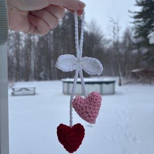 May include: A handmade crochet ornament featuring two hearts, one red and one pink, suspended from a white crocheted bow. The ornament is held up against a snowy outdoor backdrop.