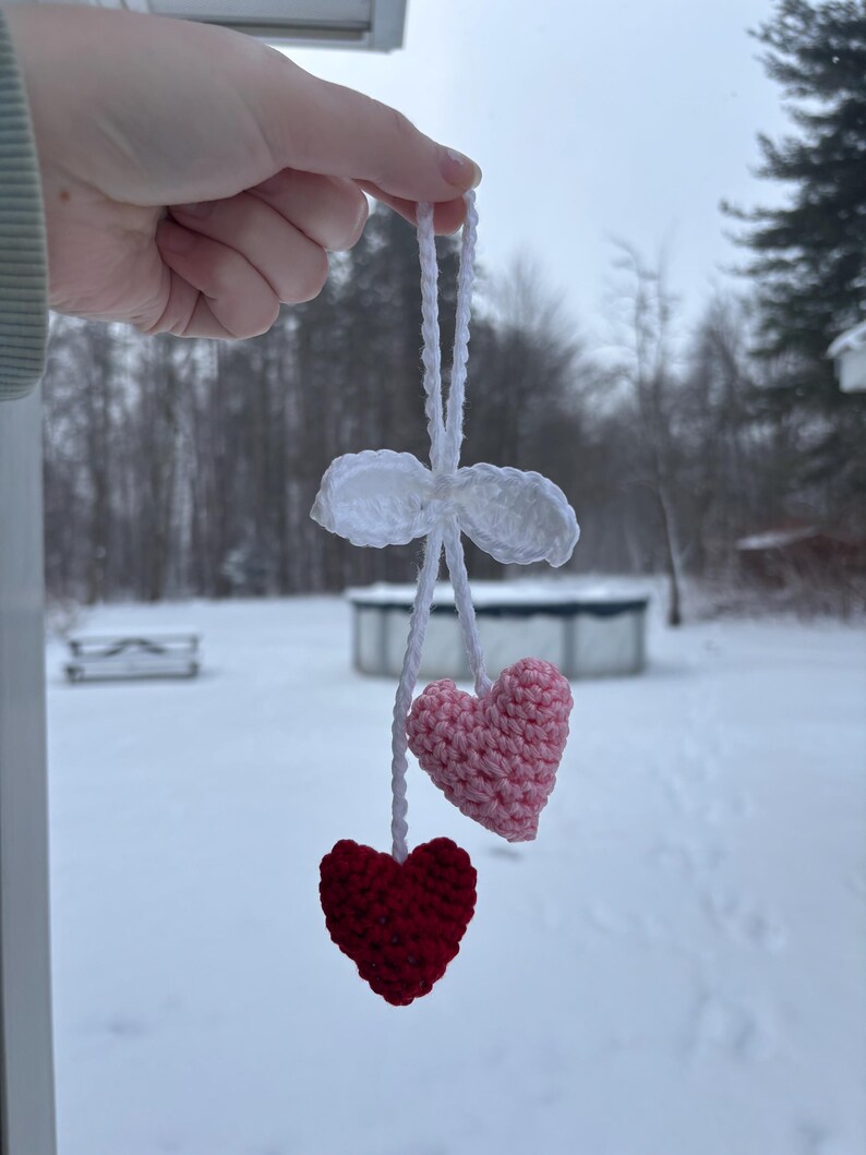 May include: A handmade crochet ornament featuring two hearts, one red and one pink, suspended from a white crocheted bow. The ornament is held up against a snowy outdoor backdrop.