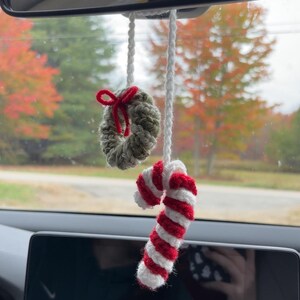 May include: Crocheted Christmas car charm with a small wreath and red bow, plus a red and white striped candy cane. The ornaments hang from a white cord, ideal for festive decoration.