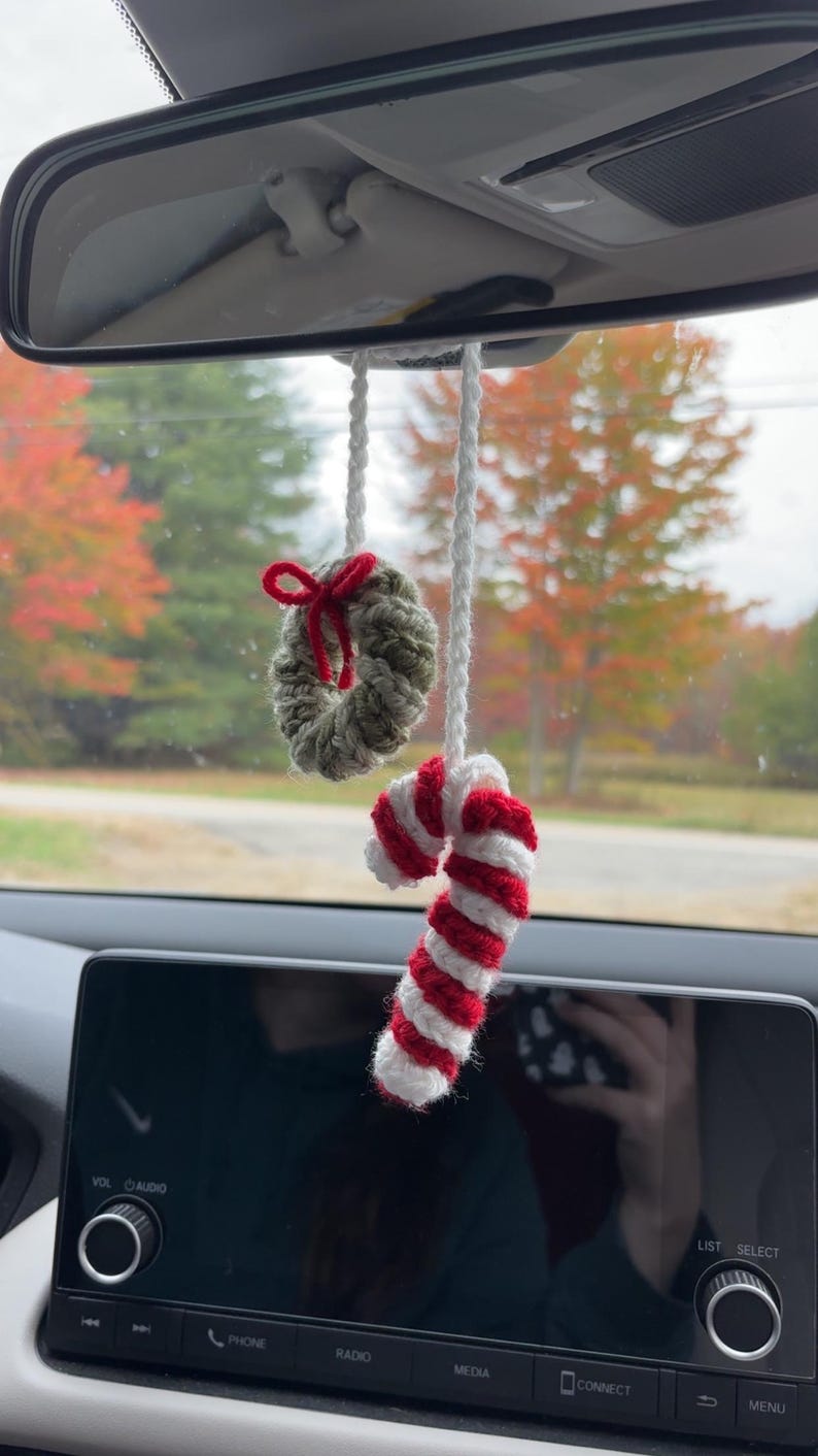 May include: Crocheted Christmas car charm with a small wreath and red bow, plus a red and white striped candy cane. The ornaments hang from a white cord, ideal for festive decoration.