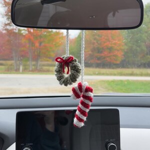 May include: Crocheted Christmas car charm featuring a small wreath with a red bow and a candy cane with red and white stripes. The ornaments hang from a white string, suspended from a car's rearview mirror.