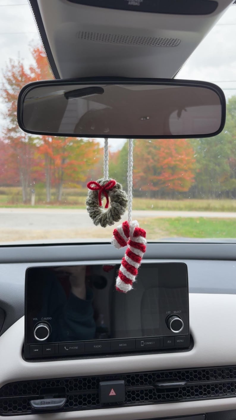 May include: Crocheted Christmas car charm featuring a small wreath with a red bow and a candy cane with red and white stripes. The ornaments hang from a white string, suspended from a car's rearview mirror.