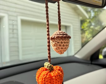 May include: A crochet car accessory featuring a small orange pumpkin with a green stem and a brown acorn, both suspended from a brown braided cord. The items are crafted with visible yarn stitches and hang together as a single decorative piece.