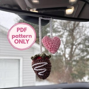 May include: A crochet car charm with a chocolate-covered strawberry and a pink heart. The strawberry is brown with white drizzle and a green top. The heart and strawberry hang from a white string. The image includes the text "PDF pattern ONLY".