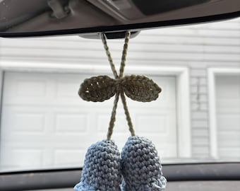 May include: A handmade car charm with two light blue crocheted bell ornaments, suspended from a beige crocheted bow. The charm hangs from a car's rearview mirror, adding a touch of handcrafted charm to the vehicle's interior.
