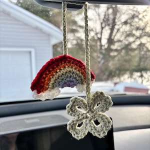 May include: A handmade crochet car charm featuring a rainbow and a four-leaf clover. The rainbow has red, orange, yellow, green, and purple stripes with white clouds. The clover is light green and hangs below the rainbow.