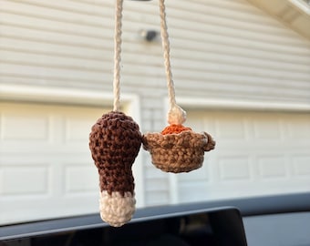 May include: A crochet car accessory hanging from a rearview mirror, featuring a brown and white chicken drumstick and a small brown pie with an orange filling and a white dollop of cream, both suspended by white crocheted strings.