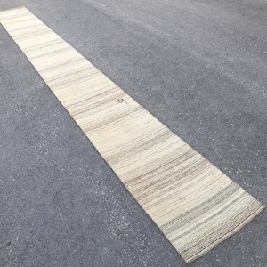 Puede incluir: Una alfombra larga tejida de color blanco roto con un sutil patrón a rayas. La alfombra está colocada sobre una superficie de asfalto gris.