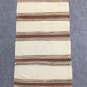 May include: A white woven rug with brown and black stripes. The rug has a fringe border.