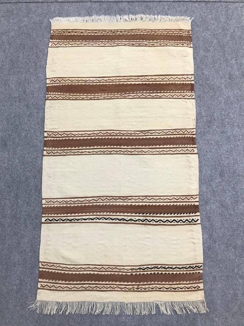 May include: A white woven rug with brown and black stripes. The rug has a fringe border.