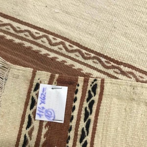 May include: A white and brown woven rug with black accents. The rug has a fringe on one side and a handwritten tag that says "114 X 202".