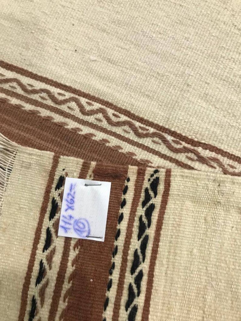 May include: A white and brown woven rug with black accents. The rug has a fringe on one side and a handwritten tag that says "114 X 202".