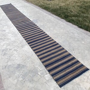 Pode incluir: Um tapete corredor longo e estreito com listras alternadas de azul marinho e bege. O tapete está colocado em um pátio de concreto cinza.