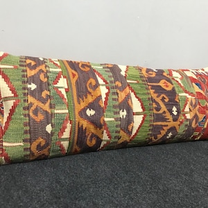 May include: A long, rectangular decorative pillow with a colorful, geometric pattern. The pillow features a mix of red, green, brown, and orange hues. The design appears to be woven, with a textured surface. The pillow is likely used for home decor.