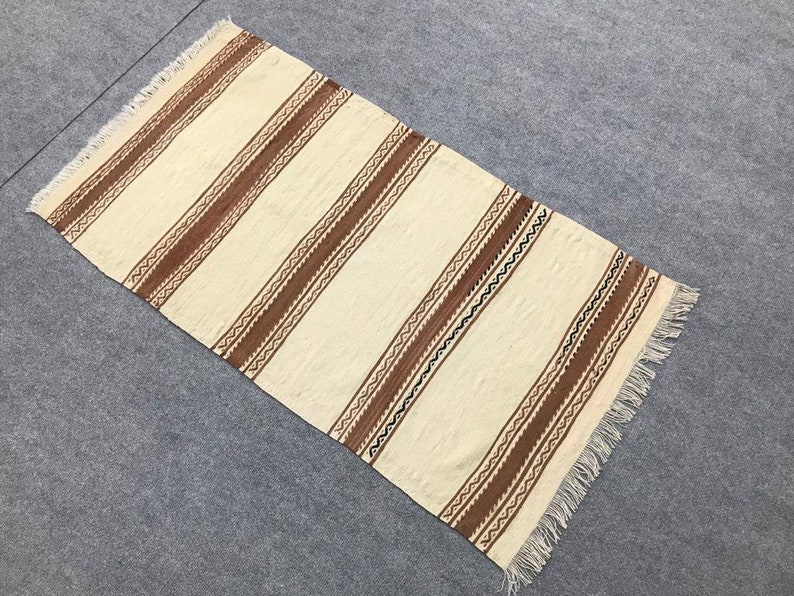 May include: A white woven rug with brown and black geometric patterns. The rug has a fringe border.