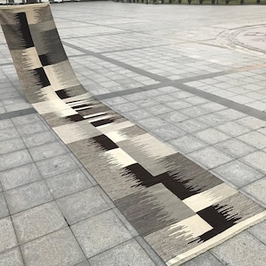 Op de afbeelding: Een lang, rechthoekig tapijt met een geometrisch patroon in grijstinten, zwart en wit. Het tapijt is dubbelgevouwen en ligt op een grijze tegelvloer.