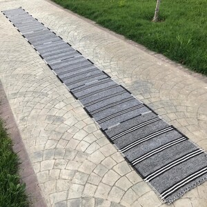 Könnte beinhalten: Ein langer, rechteckiger grauer Teppich mit horizontalen weißen Streifen. Der Teppich besteht aus gewebtem Material und hat einen schwarzen Rand. Der Teppich liegt auf einem Steinweg mit Gras auf beiden Seiten.