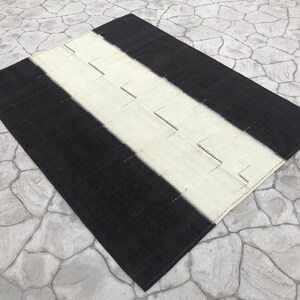 Könnte beinhalten: Ein rechteckiger Teppich mit einem auffälligen Design. Der Teppich hat einen zentralen weißen Streifen mit Zickzackmuster, der von schwarzen Streifen begrenzt wird. Der Teppich besteht aus einem gewebten Material.