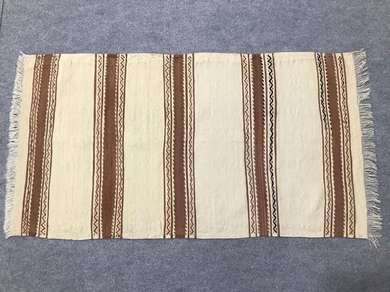 May include: A white woven rug with brown and black stripes. The rug has a fringe on all sides.
