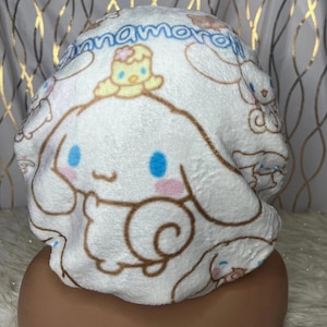 May include: White plush cap featuring a cartoon character with blue eyes, pink cheeks, and brown accents. The cap has the word "Cinnamoroll" in blue script. A small yellow character sits on top of the cap.